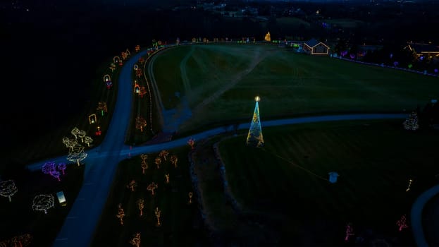 Aerial Nighttime View Of A Large Lit Christmas Tree And Multiple Smaller Trees Decorated With Colorful Lights Along A Winding Road.