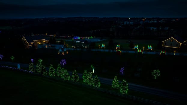 Elevated View Of A Festive Display With Two Houses Adorned In Christmas Lights And Surrounding Trees Decoratively Lit In A Rural Setting At Dusk.