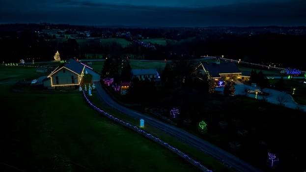 Nightfall Scene Over A Countryside Christmas Light Display With Illuminated Buildings And Trees Along A Curved Pathway.