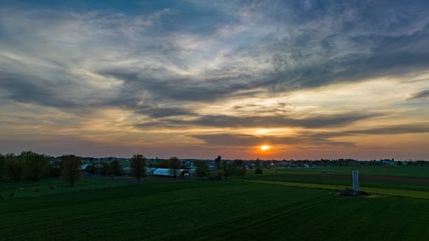 Aerial View of Dramatic Sunset Casting A Warm Glow Over A Rural Landscape With Silhouetted Trees And Buildings Under A Cloud-Streaked Sky.