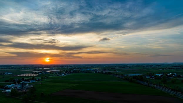 Aerial View of Dramatic Sunset Casting A Warm Glow Over A Rural Landscape With Silhouetted Trees And Buildings Under A Cloud-Streaked Sky.