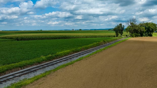 Agricultural Landscape Featuring Parallel Tracks And A Dirt Road Beside Cultivated Fields Of Varied Greens Under A Sky With Scattered Cumulus Clouds.
