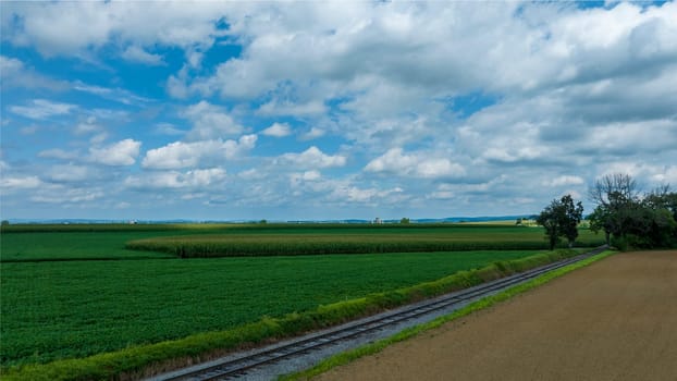 Railroad Tracks Curve Gently Through A Lush Agricultural Landscape With Fields Of Various Shades Of Green Under A Wide Sky With Broken Clouds.