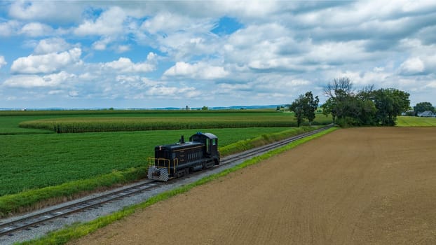 Single Locomotive Traversing Along Railway Tracks Bordering A Plowed Field And Green Crops Under A Vast Sky With Puffy Clouds.