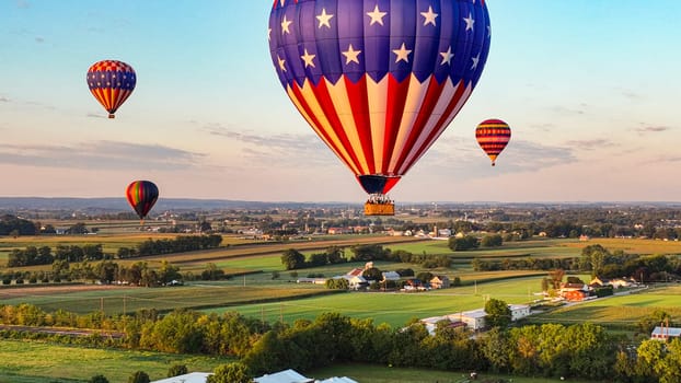 Patriotic-Themed Hot Air Balloon Dominates The Foreground With Others Dotting The Sky Above A Serene Rural Town As The Sun Sets.