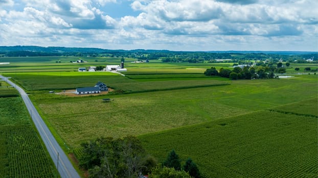 Overhead View Of A Curving Road Cutting Through Varied Agricultural Fields With A Standalone House And Adjacent Construction, Set Against A Backdrop Of Distant Trees And Cloud-Filled Sky.