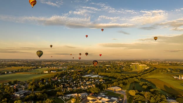 Aerial View of Sunlit Sky Filled With Hot Air Balloons Over A Tranquil Town Amidst Green Fields During An Early Morning Flight.