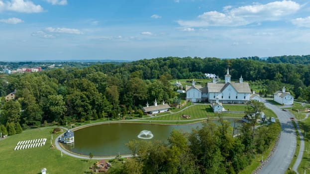 Expansive Aerial View Over A Religious Complex With Multiple White Buildings And Shining Golden Domes, A Pond With A Fountain, Set In A Verdant Landscape With A Large American Flag, With Fluffy Clouds