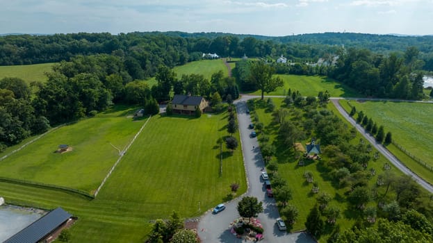 Aerial View Of A Lush Green Estate With Neatly Trimmed Lawns, A Winding Road, Fenced Paddocks, And Several Buildings Amidst A Backdrop Of Dense Trees And Far-Off White Structures Under A Clear Sky.