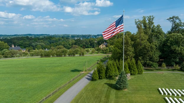 Expansive Green Field Edged By A Winding Path And Wooden Fences, With A Prominent American Flag Flying High, Overlooking A Rural Landscape Dotted With Buildings And Trees Under Scattered Clouds.