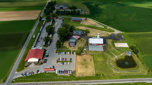 Overhead View Of A Bustling Country Market With A Red Roof, Adjacent To A Farm, Parking Lot Full Of Cars, Railroad Tracks, And Surrounding Green Fields.