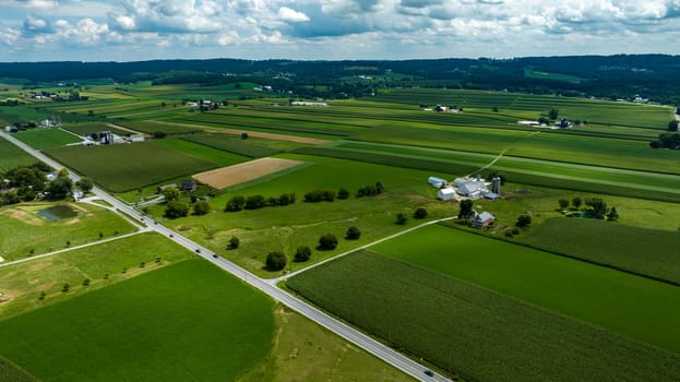 Aerial Perspective Of A Crossroads In A Verdant Rural Landscape With Patchwork Fields, Farm Structures, And A Pond Under Partly Cloudy Skies.