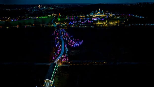 Aerial Perspective Of A Nighttime Road Decorated With Festive Lights Stretching Towards An Illuminated Complex And Reflective Water Body.
