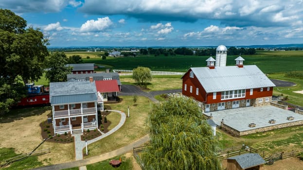 Elevated View Of A Traditional Red Barn And Outbuildings With A Stone Foundation On A Farm, Complete With A Weeping Willow And Manicured Lawns, Overlooking Expansive Agricultural Fields Under A Dynamic Sky.