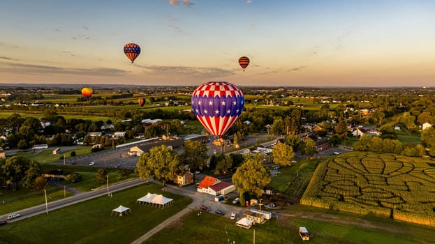 Patriotic-Themed Hot Air Balloon Dominates The Foreground With Others Dotting The Sky Above A Serene Rural Town As The Sun Sets.