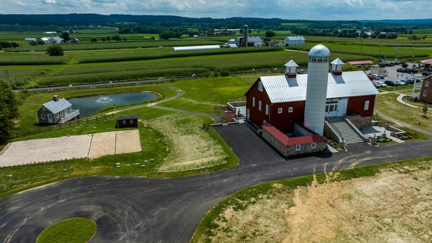 Overlooking A Farmstead Featuring A Majestic Red Barn With Silo, An Adjacent Pond With Geese, Winding Asphalt Paths, And Lush Green Crop Fields Under A Blue Sky With White Clouds.