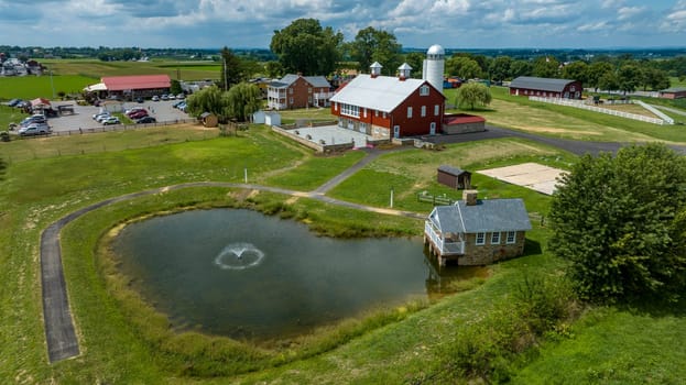 Aerial View Of A Farm Complex With Red Barns, A Large Pond With A Fountain, A Stone House, And Winding Paths, Set In A Verdant Landscape With A Parking Area Full Of Cars.