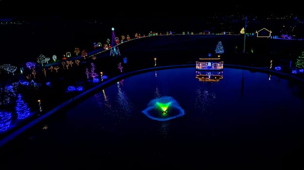 Aerial Nighttime Image Of A Pond With A Fountain And Surrounding Colorfully Lit Christmas Trees And Pathways In A Festive Setting.