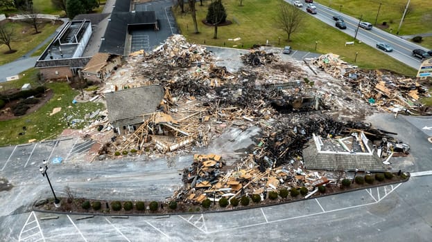 An Aerial View Of A Devastated Building Amidst Intact Structures With Cars On The Road And Green Fields In The Background Under A Cloudy Sky.