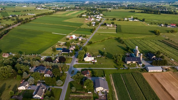 High-Angle View Of A Rural Community With Farms, Crops In Neat Rows, Roads Crisscrossing The Landscape, And Buildings Clustered Along The Thoroughfare.