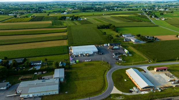 Bird's-Eye View Of Industrial Buildings Alongside A Railroad Track, Curved Roads, And Vast Agricultural Fields In A Rural Landscape At Dusk.