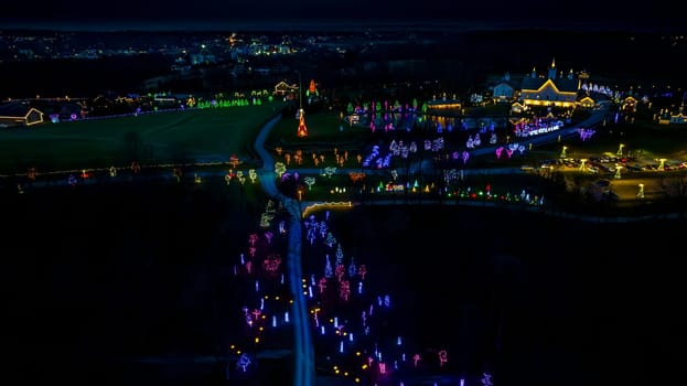 High-Angle Night View Of A Lively Holiday Lighting Display With A Road Leading To A Central Pond And Clustered Lit Structures.