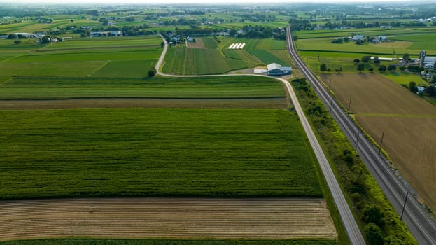High Angle View Showcasing A Stretch Of Road And Railroad Tracks Cutting Through A Lush Tapestry Of Agricultural Fields With Farm Buildings In The Distance.