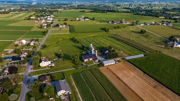 High-Angle View Of A Rural Community With Farms, Crops In Neat Rows, Roads Crisscrossing The Landscape, And Buildings Clustered Along The Thoroughfare.