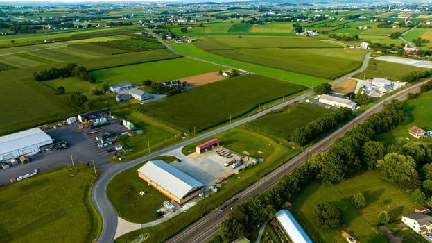 Bird's-Eye View Of Industrial Buildings Alongside A Railroad Track, Curved Roads, And Vast Agricultural Fields In A Rural Landscape At Dusk.