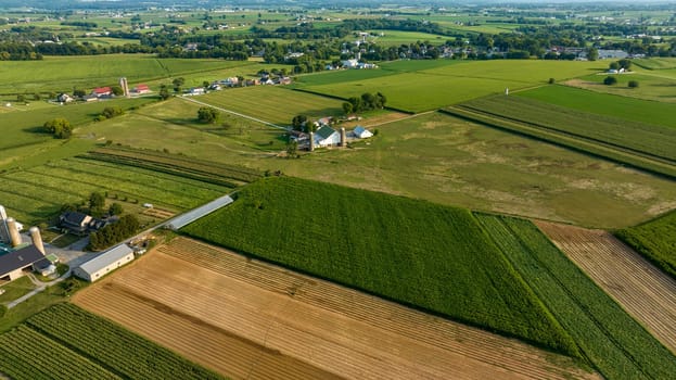 Diagonal Aerial View Of Varied Farmland With Different Crops, Farm Buildings, Silos, And A Network Of Roads In Early Evening Light.