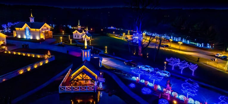 Aerial Evening Shot Of A Lit Complex With A Prominent Building, Walkways, And An Array Of Christmas Trees, Beside A Road With A Large Lit Snowflake Decoration.