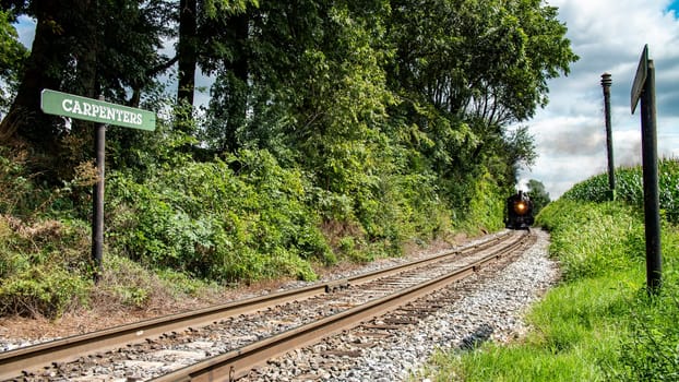 Vintage Steam Train Approaching On Tracks By The 'Carpenters' Signpost Amidst Lush Greenery On A Sunny Day.