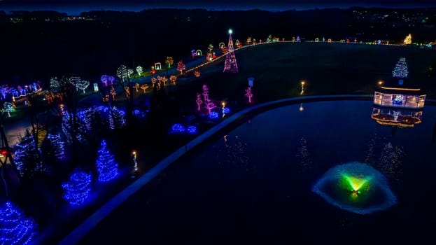 Overhead Nighttime View Of A Serene Pond With A Lit Fountain, Surrounded By A Curved Pathway With Festive Trees In Multicolored Lights.