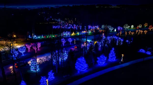 Night View Of A Vibrant Display Of Blue And Purple Christmas Lights On Trees Lining A Road With Cars And A Distant Area Aglow.