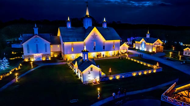 Aerial Evening View Of A Lit Central Building With Surrounding Outbuildings And Pathways, All Adorned With Festive Lighting And Trees.