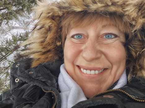 Happy, cheerful middle-aged woman in a warm jacket and a hood with fur taking selfie in winter outdoors. Female Traveler in Alaska, Lapland, Arctic, Siberia