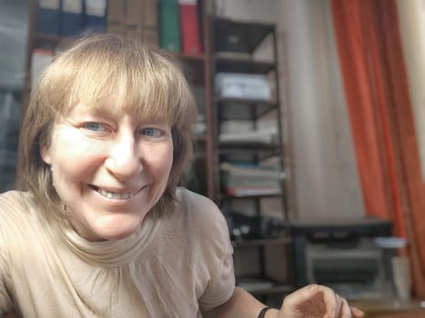 Funny blonde girl or a middle-aged woman poses for the camera with a smile and closet with folders on background. A relaxed Portrait of an Accountant, manager, blogger, freelancer in the workplace