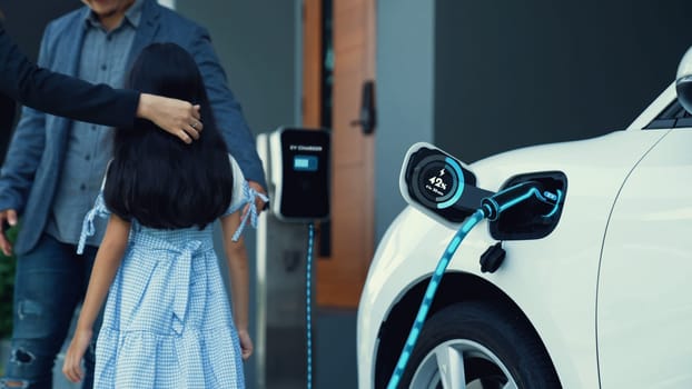 Modern family with young girl recharge electric car, EV charger from home charging station plugged in EV car in house garage. Smart and futuristic home energy infrastructure. Peruse