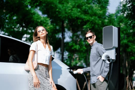 Young couple travel with EV electric car charging in green sustainable city outdoor garden in summer shows urban sustainability lifestyle by green clean rechargeable energy of electric vehicle innards