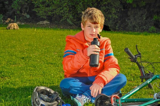 The boy sitting on the grass in a park near the bike and drinking water from a bottle