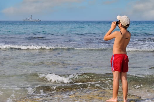 A boy stands in the water on the shores of the Mediterranean Sea and looks at a military patrol boat with binoculars.