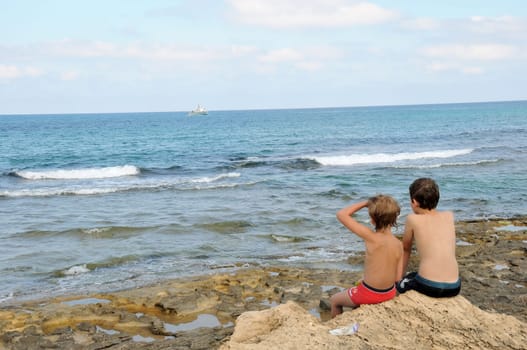 Two boys are sitting on coastal rocks, looking at a floating border boat
