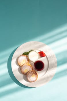 Four cheese pancakes with sour cream and strawberry jam on a white plate on a blue background. A delicious and healthy breakfast or dessert. Perfect for a food blog or recipe website.