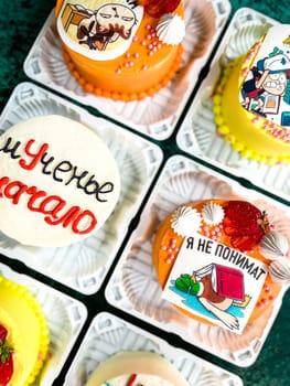 An assortment of six small square cakes with unique icing designs and decorations in plastic containers on a green surface, featuring cartoon characters, strawberries, and other decorative elements.