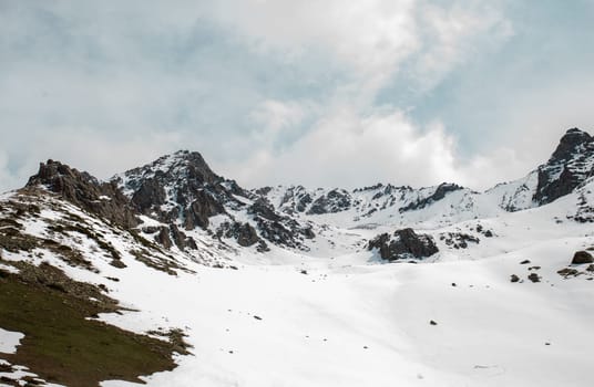 A winter landscape of snow-covered mountains and hills with a rocky summit in the distance and snow-covered hills in the foreground, taken on a bright day with a blue sky and white clouds.