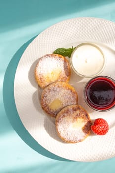 Four cheese pancakes with sour cream and strawberry jam on a white plate on a blue background. A delicious and healthy breakfast or dessert. Perfect for a food blog or recipe website.