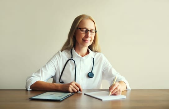 Caucasian middle-aged woman doctor with stethoscope, paper documents and digital tablet computer sitting at desk in clinic office