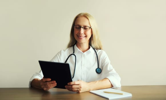 Caucasian middle-aged woman doctor with stethoscope, paper documents and digital tablet computer sitting at desk in clinic office