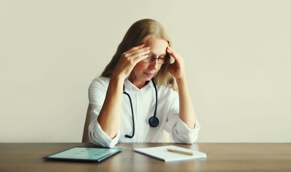 Tired caucasian middle-aged woman doctor working with digital tablet and paper documents while sitting at desk in medical clinic office