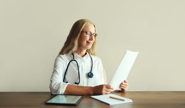 Caucasian middle-aged woman doctor with stethoscope, paper documents and digital tablet computer sitting at desk in clinic office